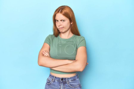 Photo for Redhead young woman on blue background frowning face in displeasure, keeps arms folded. - Royalty Free Image