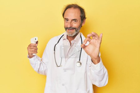 Téléchargez les photos : Médecin tenant thermomètre infrarouge en studio joyeux et confiant montrant geste ok. - en image libre de droit