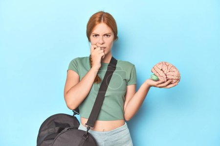 Photo for Sporty redhead with brain model on blue background biting fingernails, nervous and very anxious. - Royalty Free Image
