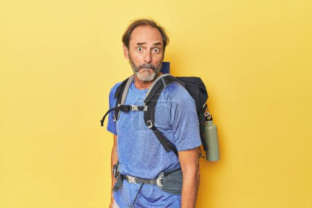 Foto de Senderista de mediana edad con mochila en estudio se encoge de hombros y los ojos abiertos confundido. - Imagen libre de derechos