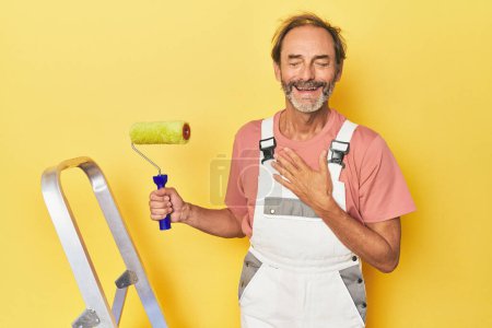 Foto de Hombre pintura amarillo telón de fondo en el estudio se ríe en voz alta mantener la mano en el pecho. - Imagen libre de derechos