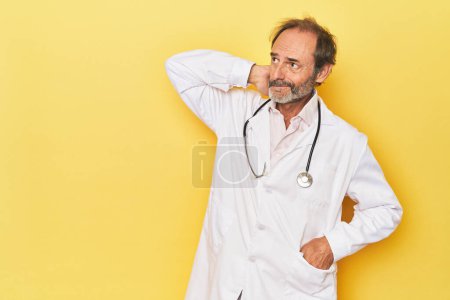 Foto de Doctor con estetoscopio en estudio amarillo tocando la parte posterior de la cabeza, pensando y haciendo una elección. - Imagen libre de derechos