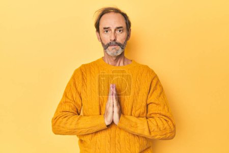 Photo for Caucasian middle-aged man in yellow studio praying, showing devotion, religious person looking for divine inspiration. - Royalty Free Image