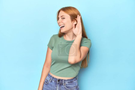Photo for Redhead young woman on blue background trying to listening a gossip. - Royalty Free Image
