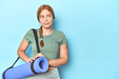 Téléchargez les photos : Rousse jeune femme tenant tapis de yoga en studio rêvant d'atteindre des objectifs et des buts - en image libre de droit