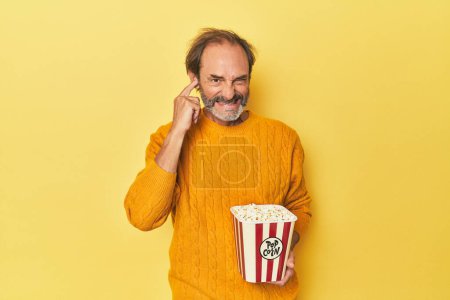 Téléchargez les photos : Homme appréciant le pop-corn en studio jaune couvrant les oreilles avec les mains. - en image libre de droit