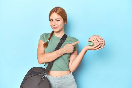 Photo for Sporty redhead with brain model on blue background smiling and pointing aside, showing something at blank space. - Royalty Free Image