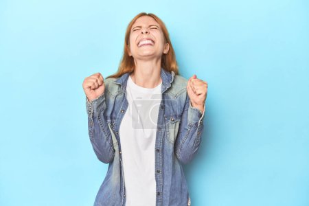 Téléchargez les photos : Jeune femme rousse sur fond bleu célébrant une victoire, passion et enthousiasme, expression heureuse. - en image libre de droit