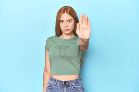 Foto de Pelirroja mujer joven sobre fondo azul de pie con la mano extendida mostrando señal de stop, que le impide. - Imagen libre de derechos