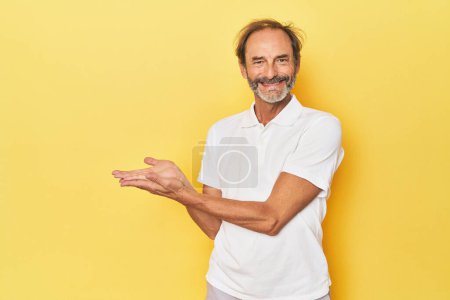 Téléchargez les photos : Blanc d'âge moyen homme en studio jaune tenant un espace de copie sur une paume. - en image libre de droit