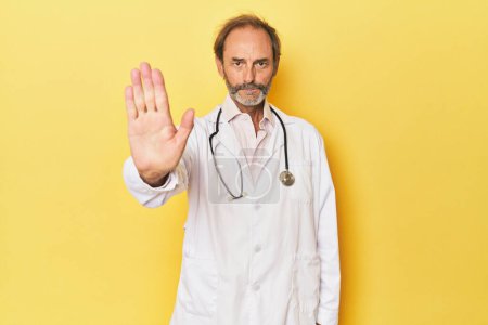 Téléchargez les photos : Médecin avec stéthoscope en studio jaune debout avec la main tendue montrant le panneau d'arrêt, vous empêchant. - en image libre de droit