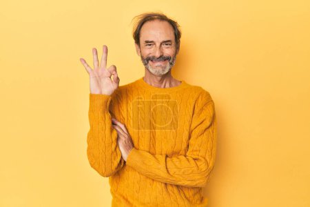 Téléchargez les photos : Caucasien d'âge moyen en jaune studio claque un oeil et tient un bon geste avec la main. - en image libre de droit