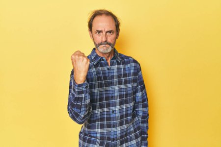 Foto de Caucásico hombre de mediana edad en estudio amarillo mostrando puño a cámara, expresión facial agresiva. - Imagen libre de derechos