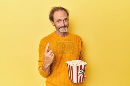 Téléchargez les photos : Homme appréciant pop-corn en studio jaune pointant du doigt vers vous comme si invitant venir plus près. - en image libre de droit