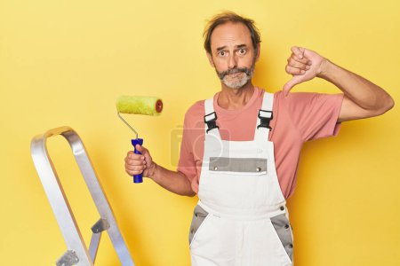 Photo for Man painting yellow backdrop in studio showing a dislike gesture, thumbs down. Disagreement concept. - Royalty Free Image