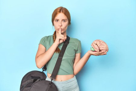 Photo for Sporty redhead with brain model on blue background keeping a secret or asking for silence. - Royalty Free Image