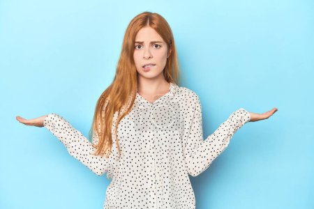 Photo for Redhead young woman on blue background confused and doubtful shrugging shoulders to hold a copy space. - Royalty Free Image