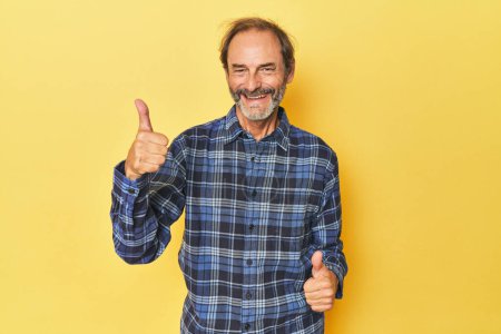 Foto de Caucásico hombre de mediana edad en estudio amarillo levantando ambos pulgares hacia arriba, sonriendo y confiado. - Imagen libre de derechos