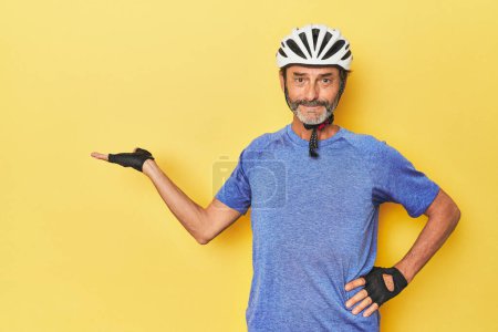 Téléchargez les photos : Cycliste portant un casque dans un studio jaune montrant un espace de copie sur une paume et tenant une autre main à la taille. - en image libre de droit