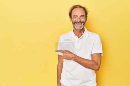 Téléchargez les photos : Homme d'âge moyen caucasien en studio jaune souriant et pointant du doigt, montrant quelque chose à blanc. - en image libre de droit