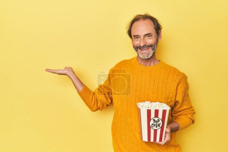 Téléchargez les photos : Homme appréciant pop-corn en studio jaune montrant un espace de copie sur une paume et tenant une autre main sur la taille. - en image libre de droit