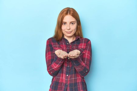 Photo for Redhead young woman on blue background holding something with palms, offering to camera. - Royalty Free Image