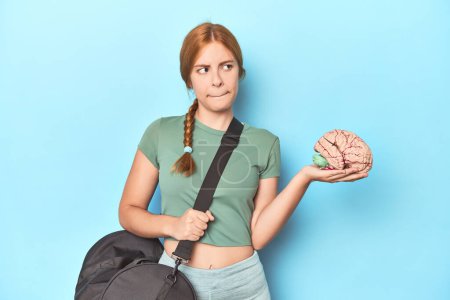 Photo for Sporty redhead with brain model on blue background confused, feels doubtful and unsure. - Royalty Free Image