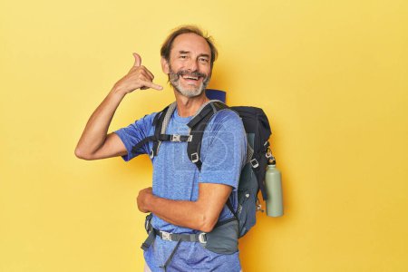 Téléchargez les photos : Randonneur d'âge moyen avec sac à dos en studio montrant un geste d'appel téléphonique mobile avec les doigts. - en image libre de droit