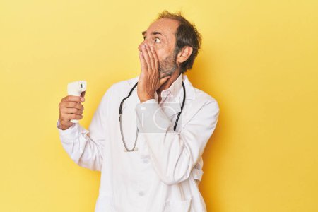 Foto de Doctor sosteniendo termómetro infrarrojo en estudio gritando y sosteniendo la palma cerca de la boca abierta. - Imagen libre de derechos