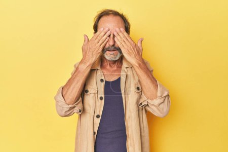 Foto de Caucásico de mediana edad hombre en amarillo estudio miedo cubriendo los ojos con las manos. - Imagen libre de derechos