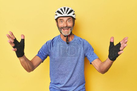 Photo for Cyclist wearing helmet in yellow studio receiving a pleasant surprise, excited and raising hands. - Royalty Free Image