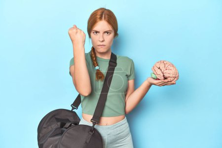 Photo for Sporty redhead with brain model on blue background showing fist to camera, aggressive facial expression. - Royalty Free Image