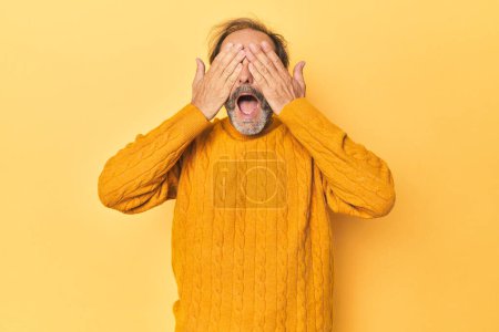 Foto de Caucásico hombre de mediana edad en estudio amarillo cubre los ojos con las manos, sonríe ampliamente esperando una sorpresa. - Imagen libre de derechos