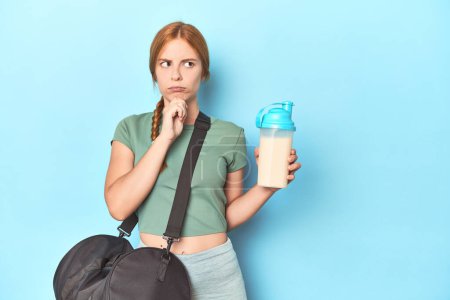 Téléchargez les photos : Redhead tenant shake protéine en studio bleu regardant latéralement avec une expression douteuse et sceptique. - en image libre de droit
