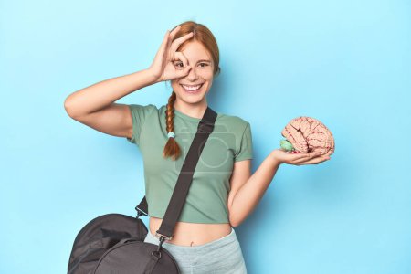 Photo for Sporty redhead with brain model on blue background excited keeping ok gesture on eye. - Royalty Free Image