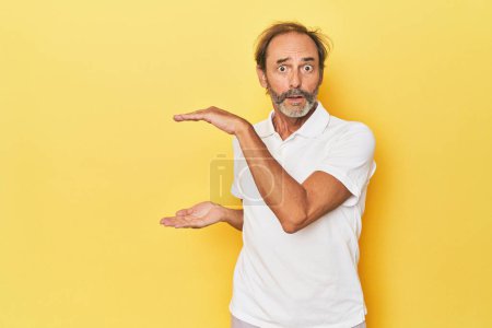 Téléchargez les photos : Blanc homme d'âge moyen en studio jaune choqué et étonné tenant un espace de copie entre les mains. - en image libre de droit