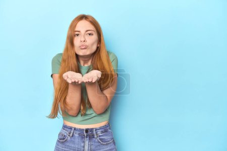 Téléchargez les photos : Rousse jeune femme sur fond bleu pliant lèvres et tenant paumes pour envoyer baiser l'air. - en image libre de droit