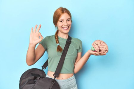 Photo for Sporty redhead with brain model on blue background cheerful and confident showing ok gesture. - Royalty Free Image