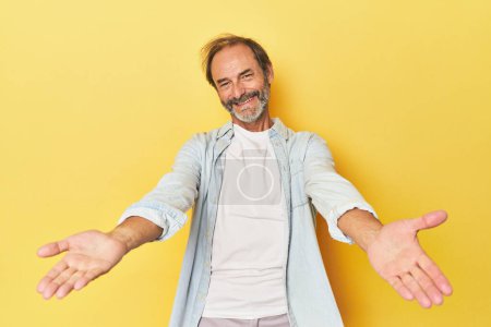 Foto de Caucásico hombre de mediana edad en estudio amarillo mostrando una expresión de bienvenida. - Imagen libre de derechos