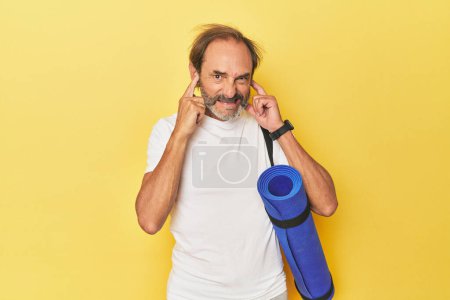 Téléchargez les photos : Homme avec tapis de yoga en studio jaune couvrant les oreilles avec les mains. - en image libre de droit