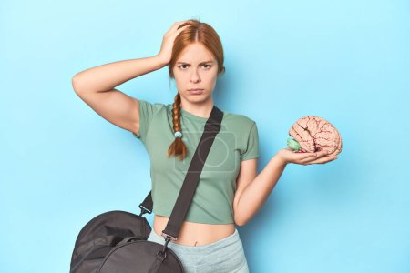 Photo for Sporty redhead with brain model on blue background being shocked, she has remembered important meeting. - Royalty Free Image