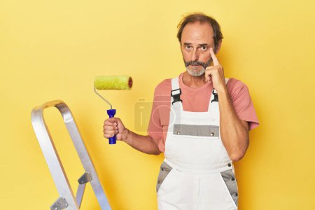 Foto de Hombre pintando amarillo telón de fondo en el estudio apuntando templo con el dedo, pensando, centrado en una tarea. - Imagen libre de derechos