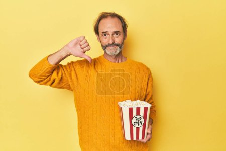 Foto de Hombre disfrutando de palomitas de maíz en el estudio amarillo mostrando un gesto de aversión, pulgares hacia abajo. Concepto de desacuerdo. - Imagen libre de derechos