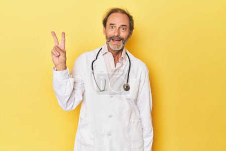 Foto de Doctor con estetoscopio en estudio amarillo alegre y despreocupado mostrando un símbolo de paz con los dedos. - Imagen libre de derechos