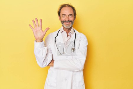 Foto de Doctor con estetoscopio en estudio amarillo sonriente alegre mostrando el número cinco con los dedos. - Imagen libre de derechos