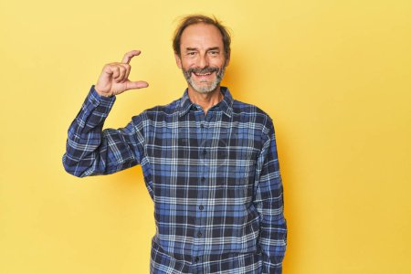 Téléchargez les photos : Blanc homme d'âge moyen en studio jaune tenant quelque chose de peu avec des index, souriant et confiant. - en image libre de droit