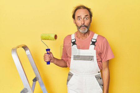 Photo for Man painting yellow backdrop in studio shrugs shoulders and open eyes confused. - Royalty Free Image