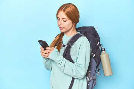 Téléchargez les photos : Roux alpiniste avec téléphone et sac à dos en studio bleu - en image libre de droit