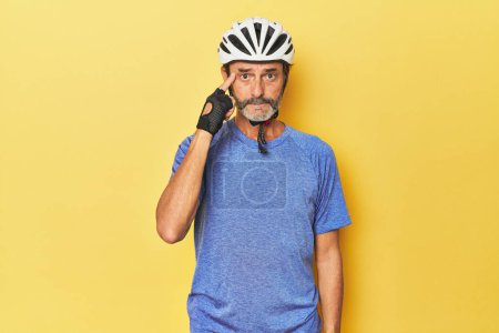 Foto de Ciclista con casco en estudio amarillo apuntando templo con el dedo, pensando, centrado en una tarea. - Imagen libre de derechos