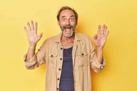 Photo for Caucasian middle-aged man in yellow studio receiving a pleasant surprise, excited and raising hands. - Royalty Free Image